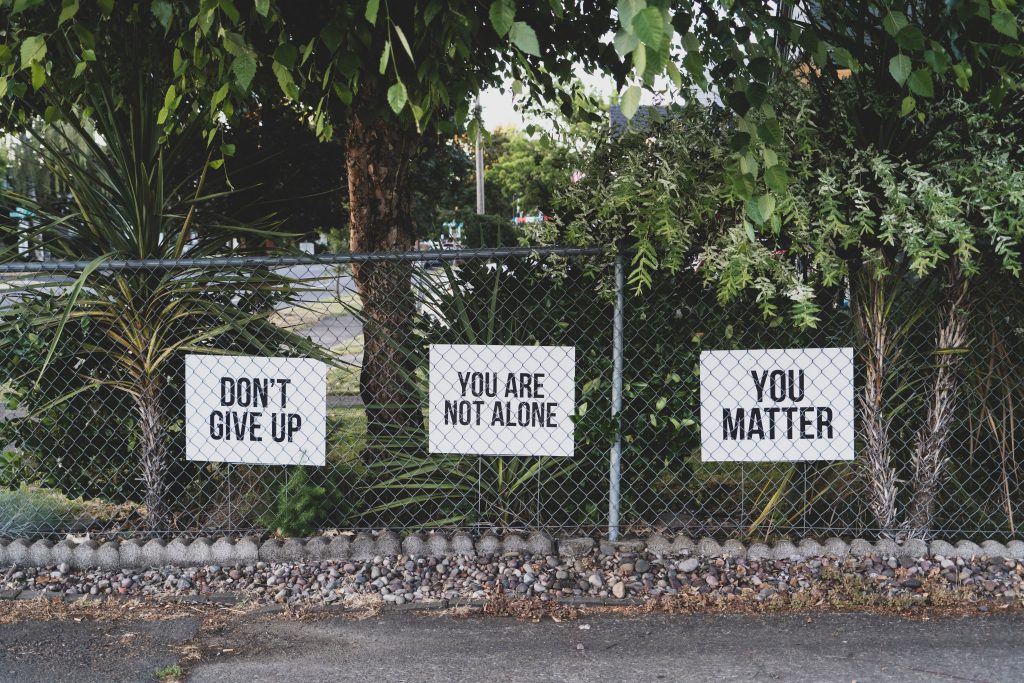 A wire fence in front of trees, with signs saying 'don't give up', 'you are not alone', and 'you matter'.
By Dan Meyers on Unsplash